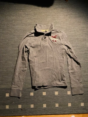 Grå henley från Hollister - Snygg grå långärmad tröja från Hollister med broderad logga och texten 'CA' i rött och vitt på bröstet. Tröjan har krage och knappar vid halsen för en avslappnad look. Perfekt till jeans eller chinos. Tröjan är i bra skick. Den är en liten L eller M skulle jag säga.