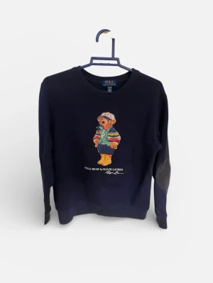 Mörkblå Polo Bear tröja Ralph Lauren - Mörkblå tröja från Ralph Lauren med ikoniska Polo Bear-trycket på bröstet. Rund halsringning och förstärkta armbågslappar. Tröjan har armbågslappar sydd på från en olycka där en del av armbågen revs, skadan syns inte på tröjan.