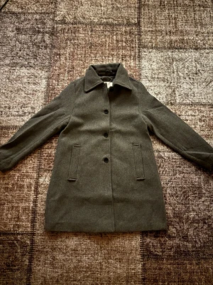 Grå klassisk ullkappa - Stilren grå kappa i ullblandning med rak passform och knäppning framtill. Kappan har två snedställda fickor och en klassisk krage. Aldrig använt! pris kan diskuteras 