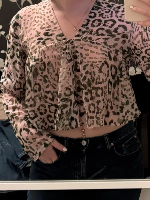 Rosa leopardmönstrad blus - Blus i ljusrosa med leopardmönster i grönt och svart. Modellen är croppad med v-ringning och långa ärmar. Tunt, lätt material. Helt ny ej använd då det inte riktigt var min stil. 