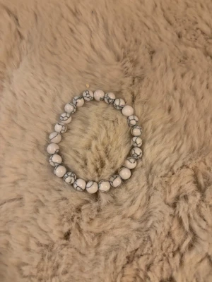 Vitt pärlarmband med marmormönster - Snyggt armband med vita pärlor och grått marmormönster. Pärlorna är runda och sitter på en elastisk tråd, vilket gör det enkelt att ta på och av. Perfekt accessoar för att lyfta din outfit med en stilren touch.