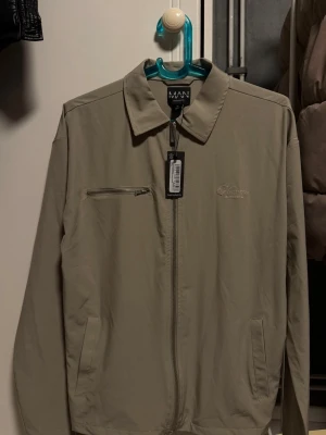 Beige jacka från MAN Official - Stilren beige jacka från MAN Official med klassisk krage och dragkedja framtill. Jackan har en bröstficka med dragkedja och två sidofickor. Materialet är tunt och passar perfekt för lager-på-lager. Diskret broderad logga på bröstet.