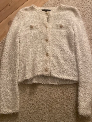 Vit fluffig kofta med pärlknappar - Supersöt vit fluffig kofta med dekorativa pärlknappar och två fejkade bröstfickor. Koftan har lång ärm och är stickad i ett mjukt, lurvigt material som känns lyxigt och trendigt. Perfekt att styla till jeans eller kjol för en chic look.