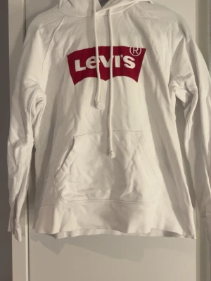 Vit hoodie från Levi's med logga - Säljer en vit hoodie från Levi's med den klassiska röda loggan på bröstet. Hoodien har huva och en stor magficka framtill. Tillverkad i mjukt bomullsmaterial och har ribbade muddar vid ärmslut och nederkant. Perfekt för en avslappnad och stilren look.