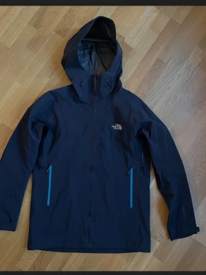 The North Face jacka - Snygg mörkblå vindjacka från The North Face med huva och dragkedja i ljusblått. Jackan har två fickor med dragkedja och justerbar huva. Perfekt för regniga dagar och outdoor-äventyr. Tillverkad i vattentätt material. Är i nyskick, knappt använd