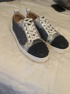 Silvriga sneakers med nitar från Louboutin - Unika sneakers från Christian Louboutin med silvriga metallicdetaljer, svart textil på sidorna och svarta nitar på tån. Skorna har vita snören och vit sula. Perfekta för dig som vill sticka ut med en edgy och lyxig stil.