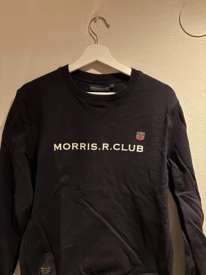 Svart sweatshirt från Morris Club - Snygg svart sweatshirt från Morris Club med tryckt text 'MORRIS.R.CLUB' på bröstet och ett litet klubbmärke. Klassisk rund halsringning och långa ärmar. Perfekt för en avslappnad och stilren look.