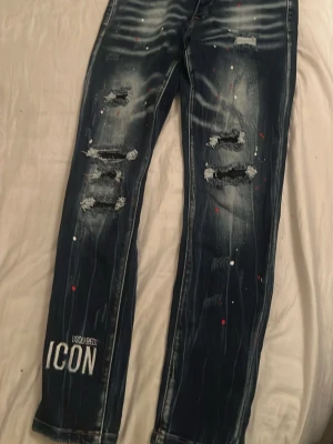 Dsquared2 ICON slitna jeans - Säljer ett par mörkblå Dsquared2 ICON jeans med slitningar, tryckta färgstänk i vitt och rött samt broderad detalj bak. Jeansen har raka ben, distressed look och tydliga slitningar framtill. Materialet är denim med stretch för skön passform.