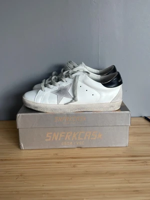 Vita Golden Goose sneakers med stjärna - Snygga vita sneakers från Golden Goose med grå stjärndetalj på sidan och svart hälparti. Skorna har klassisk låg profil, vita snören och slitna detaljer för en trendig look. Är även intresserad av byten säljer då de va förstora. Va inte blyg att fråga om byten!!! Dessa är i suveränt skick använda i gång. Pris ej hugget i sten