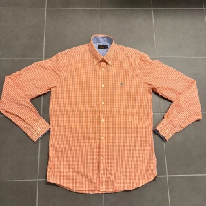 Orange rutig skjorta från Morris - Orange och vit rutig skjorta från Morris med klassisk krage och lång ärm. Skjortan har knappar framtill och en liten broderad logga på bröstet. Perfekt för en stilren och avslappnad look.