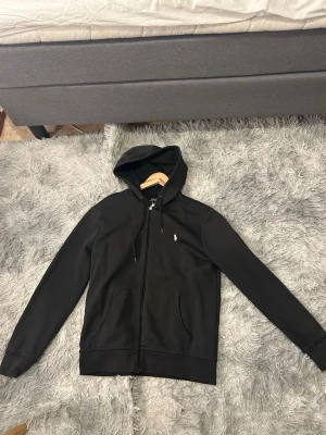 Svart hoodie från Polo Ralph Lauren - Säljer en svart hoodie från Polo Ralph Lauren med dragkedja framtill och klassisk vit logga på bröstet. Hoodien har två fickor, justerbar huva med snören och ribbade muddar. Perfekt för en chill och stilren look.