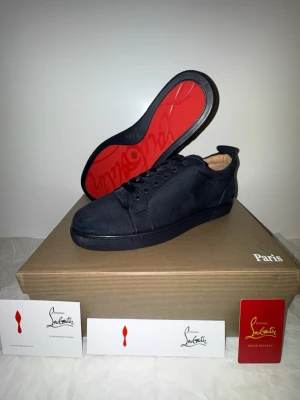 Svarta sneakers från Christian Louboutin - Stilrena svarta sneakers från Christian Louboutin i mocka med den ikoniska röda sulan. Skorna har en klassisk låg profil, svarta skosnören och diskret branding. Perfekta för dig som vill ha en lyxig och tidlös look med en touch av Paris.