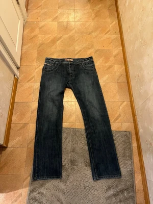 Mörkblå raka jeans från Nudie - Säljer ett par mörkblå jeans från Nudie med klassisk rak passform. Jeansen har fem fickor, diskreta slitningar och kontrastsömmar. Perfekta för dig som gillar en tidlös och avslappnad stil.