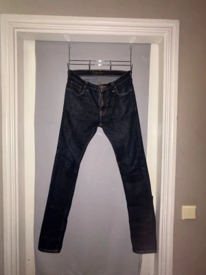 Mörkblå jeans från Nudie Jeans - Säljer ett par mörkblå jeans från Nudie Jeans med klassisk femficksdesign och raka ben. Jeansen har kontrastsömmar och läderpatch bak i midjan. Perfekta för dig som gillar stilrena och tidlösa jeans i denim.