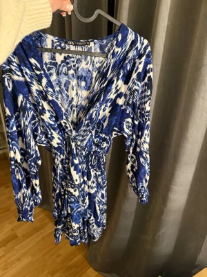 Klänning från Zara - Säljer en blåvit kortklänning från Zara med v-ringning och långa ärmar. Klänningen har ett iögonfallande abstrakt mönster och smock i midjan för en snygg passform. Perfekt för dig som gillar färgstarka plagg med unik design.