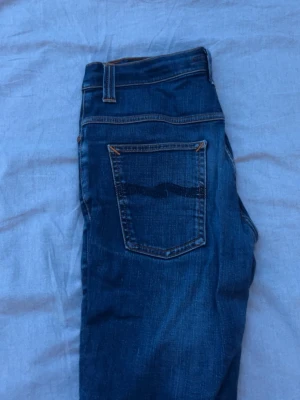 Blå jeans från nudie - Snygga blå jeans från nudie jeans med klassisk femficksdesign och raka ben. Jeansen har tydliga kontrastsömmar och en mörk tvätt som ger en stilren look. Perfekta för dig som gillar en tidlös och avslappnad stil. W. 32 L. 32 aldrig  använda, en fin liten slitning. En het favorit just nu!