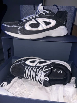 B30 Dior Countdown Sneakers - Snygga svarta Dior sneakers med stora vita CD-loggor på sidorna och vita snören. Skorna har en chunky vit sula och detaljer i mesh och syntetmaterial. Perfekta för dig som vill ha en statement sneaker med lyxig känsla och modern design.