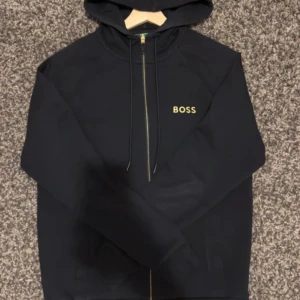 Svart hoodie från BOSS med dragkedja - Svart hoodie från BOSS med guldfärgad logga på bröstet och hel dragkedja framtill. Tröjan har huva med snörning och långa ärmar. Klassisk och stilren design, perfekt för en chill look.  Vill bli av med den så SKYNDA ER!