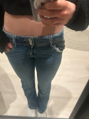 Blå bootcut jeans med dubbla knappar - Säljer ett par blå bootcut jeans med låg midja och dubbla knappar framtill. Jeansen har klassiska fem fickor och är tillverkade i denim med lätt stretch. Perfekta för dig som gillar en retro vibe och vill ha ett par jeans som sitter snyggt över höfterna. PASSAR xs eller xxs