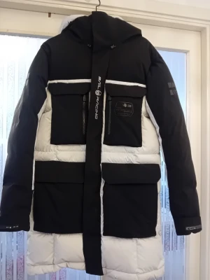 Jacka av märket Sail Racing svartvit dunjacka - Snygg svart och vit dunjacka från Sail Racing med stora fickor framtill, dragkedjor och logga på bröstet. Jackan har huva och längre modell ca 93 cm,perfekt för kalla vinterdagar. Tuff design med kontrasterande färgblock och detaljer.Det finns en lagning inuti jackan,men inget som påverkar funktionen eller utseende.  Lagningen finns på sista bilden . Storleken är L 
