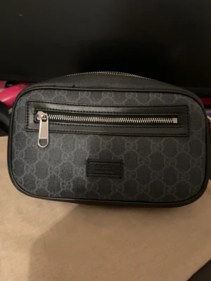 Svart axelväska från Gucci - Snygg svart axelväska från Gucci med klassiskt GG-mönster i grått. Väskan har en silverfärgad dragkedja framtill och justerbart band i rött och blått. Tillverkad i canvas och läderdetaljer. Perfekt för att bära det viktigaste med stil. Använt ett par gånger haft den max 1-2 månader 