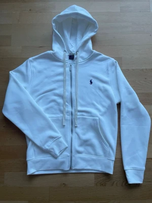 Vit hoodie från Polo Ralph Lauren - Klassisk vit hoodie från Polo Ralph Lauren med dragkedja framtill, två fickor och justerbar huva med snören. Broderad blå logga på bröstet. Tillverkad i mjuk bomullsblandning som känns skön mot huden. Perfekt för en clean och stilren look.