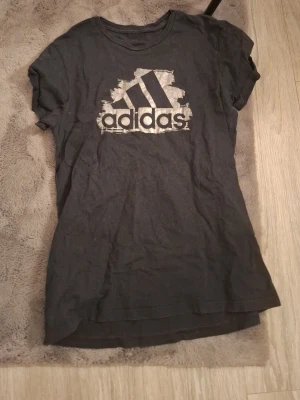 Svart Adidas t-shirt med logga - Snygg svart t-shirt från Adidas med stor vit logga framtill. Klassisk rund halsringning och korta ärmar. Perfekt för en sportig och avslappnad stil. Tillverkad i mjuk bomull för skön känsla hela dagen.