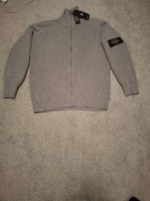 Grå ziptröja från Stone Island - Säljer en snygg grå stickad ziptröja från Stone Island med ribbad struktur och klassisk logotyp-patch på ärmen. Tröjan har hög krage, hel dragkedja och långa ärmar. Perfekt för lager-på-lager och streetwear-stil. Ny pris 4000 säljs för 900