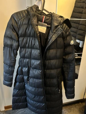 Svart lång dunjacka från Moncler - Snygg svart lång dunjacka från Moncler med quiltat mönster, huva och dragkedjor på fickorna. Jackan har en klassisk Moncler-logga på ärmen och är perfekt för kalla vinterdagar. Stilren design som håller dig varm och bekväm. Har en liten skada vid fickan som går att sy ihop 