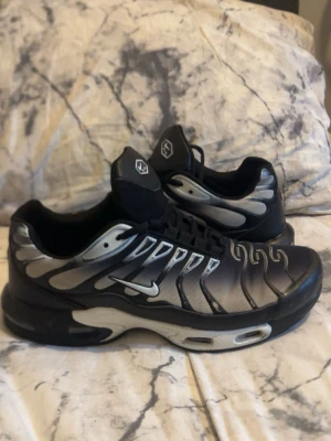 Nike Air Max Plus svarta och silver - Nike Air Max Plus sneakers i svart och silver med ikoniska vågformade detaljer och synlig Air-sula. Ovandelen är i mesh och syntetmaterial, med vita och silvriga accenter samt klassisk Swoosh-logga på sidan. Skorna har en sportig och futuristisk vibe.