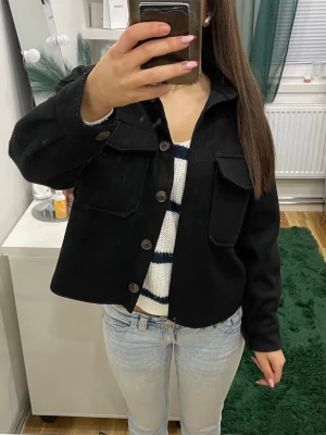 Svart kappa med stora fickor - Snygg svart tunn kappa med stora bröstfickor och knappar framtill. Modellen är boxig och croppad, vilket ger en trendig look. Materialet ser ut att vara tjockt och mjukt, perfekt för lager på lager. Passar till jeans och t-shirt för en avslappnad stil.