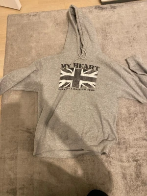 Grå my heart hoodie  - Grå hoodie med stor huva och tryck av brittiska flaggan på bröstet. Texten 'MY HEART BEATS TO A BRITISH TUNE' ger en cool vibe. Mjuk och skön, perfekt för dig som gillar streetstyle och vill sticka ut lite extra. skriv vid frågor eller funderingar 