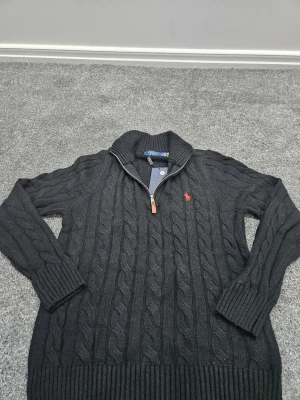 Svart kabelstickad tröja Polo Ralph Lauren - Kabelstickad svart tröja från Polo Ralph Lauren med halv dragkedja och röd broderad logga på bröstet. Tröjan har hög krage, långa ärmar och breda ribbade muddar. Perfekt för lager-på-lager och snygg till jeans.