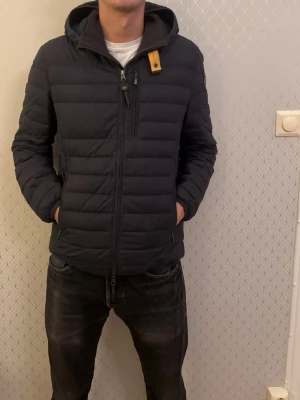 Parajumpers jacka  - Snygg svart dunjacka från Parajumpers med huva och dragkedja framtill. Jackan har en gul detalj vid halsen och logga på ärmen. Flera fickor med dragkedja och quiltad design. Perfekt för kalla vinterdagar.