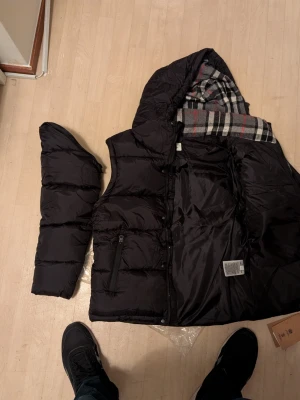 Svart dunjacka med huva från Burberry london - Säljer en svart dunjacka från Burberry med huva och dragkedja. Jackan kan bli till en väst och har storleken L