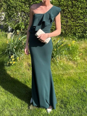 Mörkgrön balklänning med volang - Elegant mörkgrön långklänning med volangdetaljer och oneshoulder-design från Lulus. Klänningen har en figurnära passform och är perfekt för speciella tillfällen. Materialet är slätt och följsamt, vilket ger en lyxig känsla. Perfekt till studentbal!