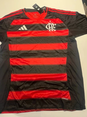 Flamengo matchtröja Adidas röd/svart - Snygg Flamengo matchtröja från Adidas i rött och svart med klassiska horisontella ränder. Tröjan har klubbens logga och Adidas-märket på bröstet samt röda detaljer på axlarna. Tillverkad i lätt och ventilerande material.