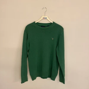 Grön stickad tröja från Gant - Mysig grön stickad tröja från Gant med rund halsringning och diskret logga broderad på bröstet. Tröjan har långa ärmar och ribbade muddar vid ärmslut och nederkant. Perfekt för lager-på-lager-stil.