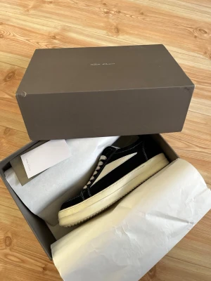 Svarta RlCK OWENS sneakers i mocka - Svarta sneakers från RlCK OWENS i mocka med vit sula och vita detaljer på sidan. Klassisk låg modell med vita skosnören och kontrastsömmar. Perfekta för dig som gillar stilrena och trendiga skor med en clean look.