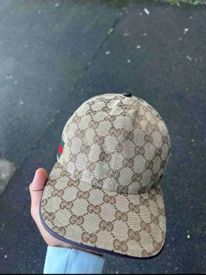Beige keps  - Säljer en beige  1.1 keps med det klassiska GG-monogrammet över hela kepsen. Kepsen har en böjd skärm och en grön-röd rand på sidan. Tillverkad i canvasmaterial och har en exklusiv streetwear-vibe.