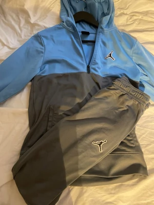 Jordan blågrå loungewear set - Snyggt loungewear set från Jordan med halvzip hoodie i ljusblått och grått samt matchande gråa byxor. Båda plaggen har den klassiska Jumpman-loggan broderad framtill. Perfekt för chill dagar hemma eller till träningen.