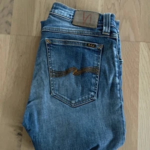 Blå jeans från Nudie Jeans - Säljer ett par klassiska blå jeans från Nudie Jeans med gul söm på bakfickan och läderpatch i midjan. Jeansen har en smal passform och är tillverkade i mjukt denimtyg. Perfekta för dig som gillar stilrena och tidlösa jeans.