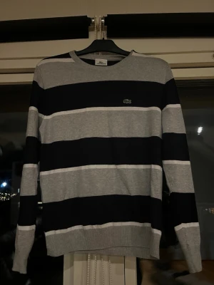 Randig tröja från Lacoste grå/svart - Snygg tröja från Lacoste med breda horisontella ränder i grått och svart. Klassisk rund halsringning och ribbade muddar. Ikoniska Lacoste-loggan med krokodil på bröstet. Perfekt för dig som gillar stilrena och sportiga plagg.