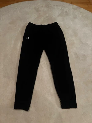 Svarta mjukisbyxor från Under Armour - Säljer ett par svarta mjukisbyxor från Under Armour med dragsko i midjan och resår vid bensluten. Byxorna har en enkel och clean look med Under Armour-logga på vänster lår. Perfekta för chill eller träning.