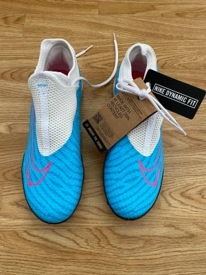 Nike fotbollsskor blå och vita - Nike fotbollsskor med Dynamic Fit, i blått och vitt med rosa detaljer. Skorna har snörning och en ovandel i syntetmaterial med mesh för bra ventilation. Sulan är svart och mönstrad för bra grepp på planen. Perfekta för dig som vill sticka ut på fotbollsplanen. Helt nya aldrig använda kommer med kartongen