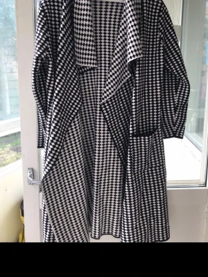 Svartvit rutig kappa med oversized passform - Snygg svartvit kappa med rutigt mönster och lös, oversized passform. Kappan har stora slag och två fickor framtill. Perfekt för dig som vill sticka ut med ett grafiskt uttryck.