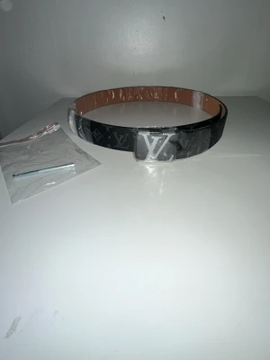 Louis Vuitto - Svart bälte från Louis Vuitto med klassiskt monogrammönster och stor L spänne i silver. Bältet har en stilren design och insida i ljusbrunt. Perfekt accessoar för att lyfta din outfit.