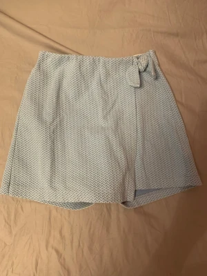 Ljusblå omlott-skorts med rosett - Supersnygga ljusblå skorts för barn med diskret zigzag-mönster och omlottdesign framtill. Skortsen har en söt rosettdetalj i midjan och elastisk baksida för extra komfort. Perfekt för dig som vill ha kjol-look men shortskomfort.