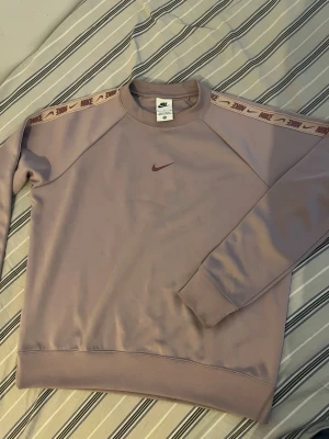 Ljuslila Nike sweatshirt med logga - Säljer en ljuslila sweatshirt från Nike med vinröd swoosh på bröstet och dekorativa band med Nike-logga längs ärmarna. Tröjan har rund hals, ribbade muddar och är tillverkad i mjukt material. Perfekt för en avslappnad och sportig stil.