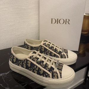 Dior sneakers med monogram och snörning - Snygga Dior sneakers med marinblått och beige monogrammönster, klassisk snörning och vit platt sula med stjärndetalj undertill. Skorna har rund tå och texten 'Christian Dior' på plösen. Tillverkade i canvas och gummisula, perfekt för dig som gillar lyxiga detaljer. Allt på bilden medföljs! Även bevis på orderbekrätelse 